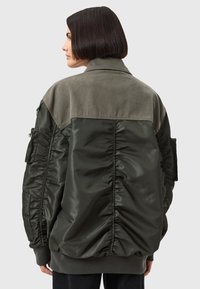 Giacca bomber verde oliva con un tessuto opaco e testurizzato nella parte superiore e una sezione inferiore lucida e imbottita. Presenta polsini a costine e tasche laterali.