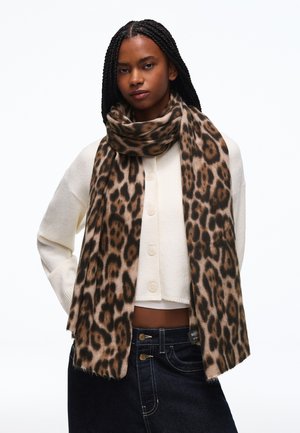 Ung kvinde med fletninger iført en hvid knappet sweater og et langt leopardmønstret tørklæde over mørke denimjeans.