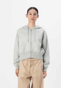 Sweat à capuche gris zippé, en matériau doux, avec une poche frontale et un ourlet côtelé, associé à un pantalon beige à taille élastique.