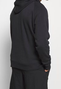 Persona che indossa un semplice hoodie nero e pantaloni neri, con la mano destra in tasca, di spalle rispetto alla fotocamera su uno sfondo bianco.
