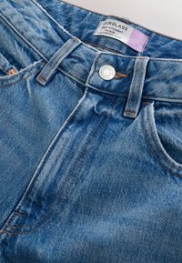 Jean denim bleu taille mi-haute, coupe droite aux chevilles, avec bouton frontal, passants pour ceinture et étiquette visible à l'intérieur de la ceinture.