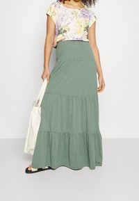 Maxi rok in olijfgroen met verschillende lagen ruches, gecombineerd met een bloemen top met korte mouwen. Soepele textuur, elastische tailleband. Model met een schoudertas.