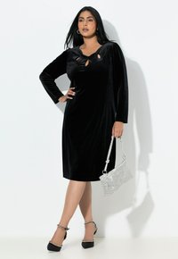 Czarna welurowa sukienka z długimi rękawami, dekoltem w kształcie łezki, o długości do kolan; uzupełniona srebrną torebką z koralików i czarnymi szpilkami.