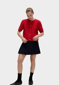 Cardigan court à manches courtes en tricot texturé rouge avec des boutons dorés, associé à une jupe plissée noire et des chaussettes noires. Lignes simples et épurées.
