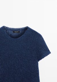 Marineblauwe gebreide trui met een zachte, pluizige textuur, korte mouwen en een ronde hals met geribbelde rand. Merklapje zichtbaar aan de binnenkant.