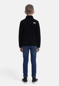 Παιδί που στέκεται γυρισμένο προς τα πίσω, φοράει μαύρο μπουφάν North Face, μπλε τζιν και μαύρα ύψους μέχρι τον αστράγαλο αθλητικά παπούτσια, σε απλό λευκό φόντο.