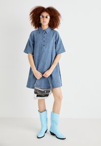 Robe courte à manches courtes texturée bleue avec un col et des boutons, associée à un sac à main en argent brillant et des bottes de cow-boy bleu clair.