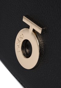 Bolso de cuero negro texturizado con un acento de hardware circular en tono dorado que lleva un logo, añadiendo contraste y detalle al diseño.
