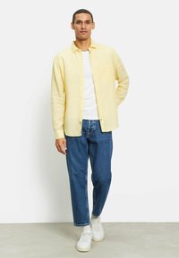 Gelbes Leinenhemd, darunter ein weißes T-Shirt, blaue Jeans und weiße Sneaker. Verfügt über Knöpfe, eine Brusttasche und einen lässigen Schnitt.