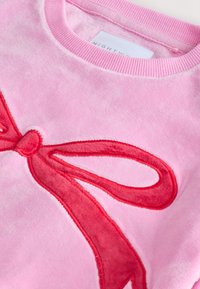 Sweat-shirt en polaire rose avec un grand motif de ruban rouge, texture douce, col côtelé et détails cousus sur le noeud.