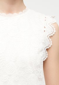 Κοντινό πλάνο λευκού μπλουζιού χωρίς μανίκια με δαντέλα, διακοσμημένο με λουλουδένια σχέδια και κυματιστές άκρες στον λαιμό και τις μασχάλες.