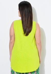 Top sin mangas de color verde lima con escote redondo, dobladillo trasero ligeramente más largo y textura suave. La modelo lleva pantalones cortos estampados.