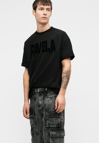 Jaunas vyras vilki juodą "FAVELA" marškinėlį ir rūgšties plovimo cargo kelnes, demonstravęs apatinį rankos tatuiruotę su laikrodžiu ir gėlių raštu.