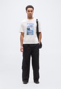 Άνδρας που φοράει λευκό T-shirt με σχέδιο ωκεάνιου κύματος και το κείμενο "HUGO", μαύρο παντελόνι cargo, μαύρα παπούτσια και μαύρη τσάντα ώμου σε λευκό φόντο.