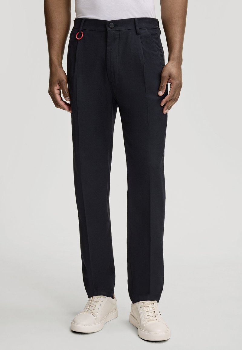 Pantalons navy à coupe droite, dotés d'une fermeture avec bouton visible et d'une boucle rouge en accent. Le matériau semble lisse et léger.