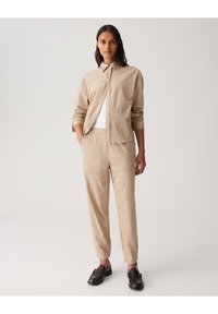 Chemise beige à manches longues avec boutons, assortie à un pantalon tapered. Tissu lisse avec des poches minimales, complété par des mocassins noirs.
