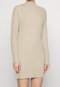 Abito a maglia beige a coste con colletto alto e maniche lunghe. Presenta motivi verticali e un design aderente, che arriva a metà coscia.