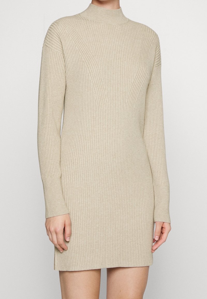 Abito a maglia beige a coste con colletto alto e maniche lunghe. Presenta motivi verticali e un design aderente, che arriva a metà coscia.