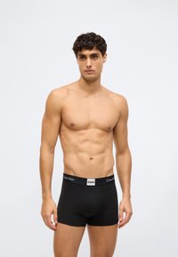 Mladý muž s krátkými kudrnatými vlasy, oblečený v černých boxerkách Calvin Klein, na jednoduchém světle šedém pozadí.