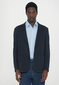 Blazer azul marino con dos bolsillos frontales, cierre de un solo botón, usado sobre una camisa de color azul claro. Corte ajustado, textura suave, diseño minimalista.