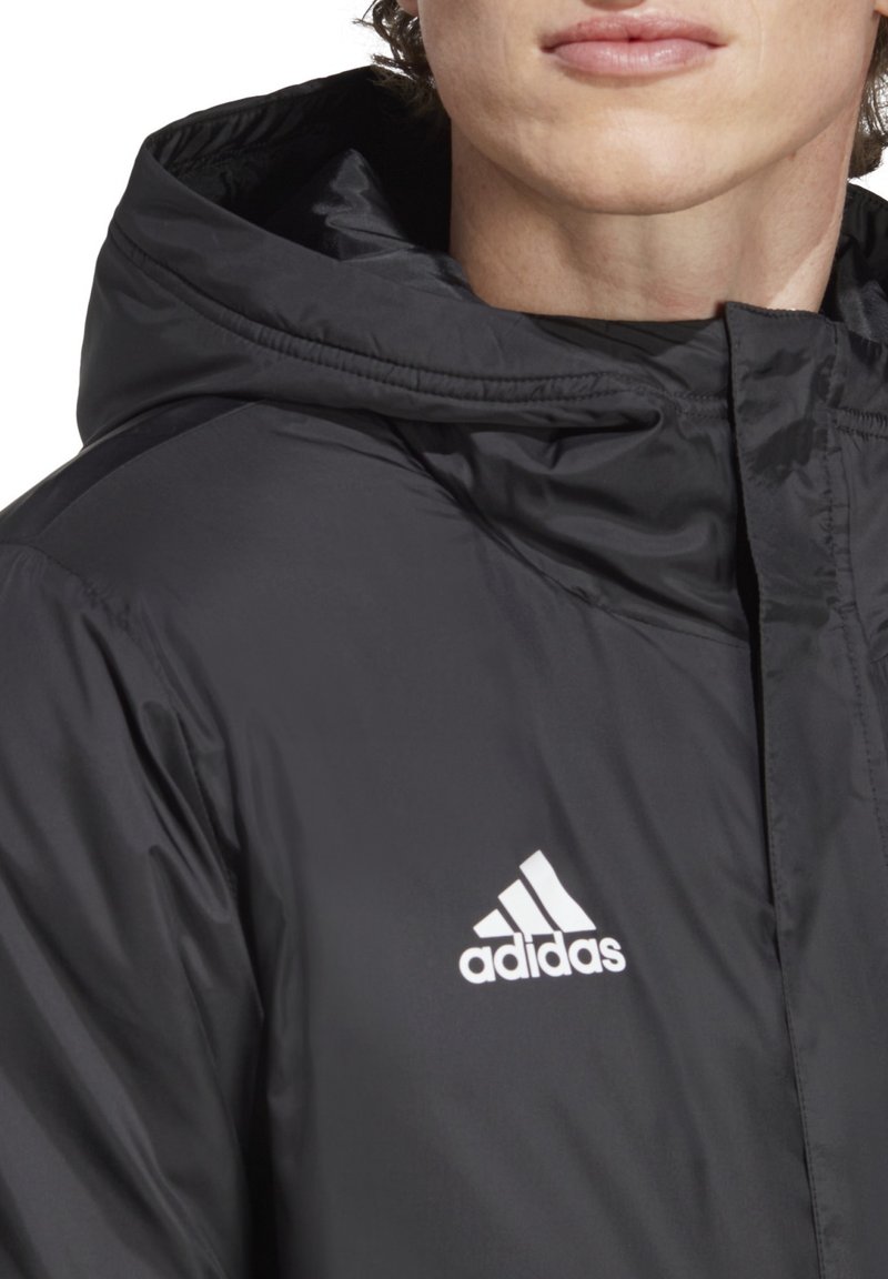 Sort polstret jakke med hætte, der har et hvidt Adidas-logo på venstre bryst. Materialet ser glat og blankt ud.