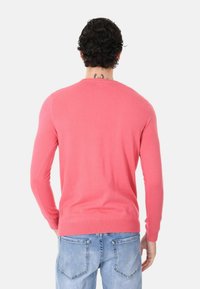 Maglione lavorato a maglia color corallo con maniche lunghe, scollo rotondo e orlo a coste. Indossato sopra jeans azzurri. Tatuaggio visibile alla nuca.