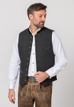 Mann trägt traditionelle bayerische Kleidung mit weißem Hemd, dunkler gemusterter Weste und braunen bestickten Lederhosen mit dekorativer Gürtelschnalle.