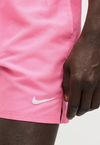 Χέρι με σκουρόχρωμο δέρμα που ακουμπά δίπλα σε ροζ σορτς Nike με λευκό λογότυπο Swoosh και μικρή πλαϊνή σχισμή, ορατά πάνω σε ανοιχτόχρωμο φόντο.