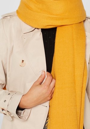 Personne portant un trench-coat beige et un pull noir, tenant d'une main un grand foulard jaune moutarde enroulé autour de son cou.