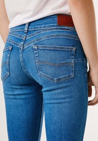 Person som har på seg tettsittende blå jeans med baklommer og en hvit skjorte som er stukket inn, synlig lavere rygg og øvre legger.