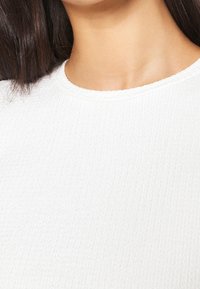 Camiseta blanca de tejido acanalado con escote redondo. La textura parece suave y presenta un patrón sutil. La vista de cerca destaca los detalles del material.