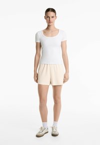 Bílý krátký rukáv top s kulatým výstřihem, spárovaný s béžovými sportovními šortkami. Model nosí béžové a bílé tenisky s bílými ponožkami.