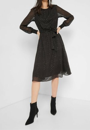 Femme portant une robe midi noire avec des manches longues transparentes, des ornements à pois, une ceinture à la taille, et des bottines à talons aiguilles pointues noires.
