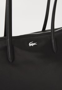 Black tote bag made of nylon with two top handles, silver zip closure, and a small white crocodile logo on the front. Smooth texture.