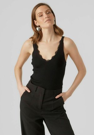 Black ribbed tank top with a deep V-neckline and scalloped lace trim, paired with high-waisted black trousers. Silver heart-shaped earrings.