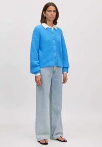 Blauwe gebreide cardigan met een textuurpatroon, knoopsluiting, oversized pasvorm, gedragen over een witte blouse met kraag en gecombineerd met lichte spijkerbroek.