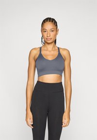 Šedá sportovní podprsenka s výstřihem do tvaru lžičky a širokými ramínky, zkombinovaná s černými legínami s vysokým pasem. Tkanina je hladká a pružná.