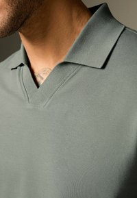 Polo shirt i dæmpet grøn med v-hals og klassisk krave. Stoffet har en tekstureret overflade, der er velegnet til afslappet brug.