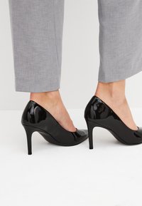 Zwarte laquéleren hoge hakken met een afgeronde neus, verhoogde naaldhak en glad oppervlak, gecombineerd met lichtgrijze op maat gemaakte broek.