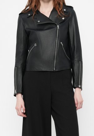 Kobieta nosząca czarną skórzaną kurtkę motocyklową z srebrnymi zamkami błyskawicznymi na przednich kieszeniach i rękawach, w połączeniu z czarnymi spodniami.