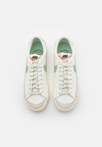 Valkoiset lenkkarit, joissa on mintunvihreät yksityiskohdat, teksturoidut harmaat mokkasinokkapäät ja beige nauhat. Sisältää oranssin ja mustan Nike-logon kielekkeellä.