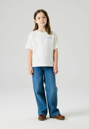 Jeune fille debout, portant un t-shirt blanc avec un texte bleu, un jean bleu ample et des chaussures marron, sur un fond clair uni.