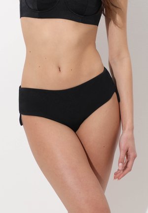 Donna che indossa slip bikini neri a vita alta e un top bikini nero abbinato, in piedi con il braccio destro rilassato lungo il corpo.