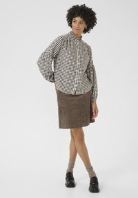 Una donna con capelli ricci indossa una camicia a scacchi bianca e nera, una gonna in cotone a coste marrone e scarpe nere con calzini beige, sorridendo leggermente.