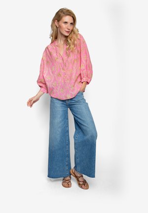 Rosa Bluse mit goldenem Palmenmuster, Puffärmeln und einer Knopfleiste, kombiniert mit weit geschnittenen blauen Jeans und braunen Sandalen.