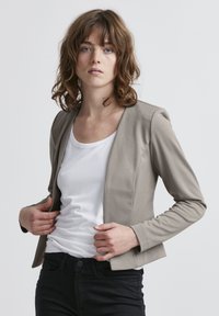 Beige sovitettu bleiseri, jossa on pyöreä kaula-aukko, pitkät hihat ja sileä kangas, yhdistetty valkoiseen paitaan ja mustiin housuihin. Minimalistinen muotoilu.