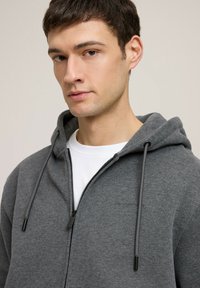 Szara bluza z kapturem na zamek, o miękkiej teksturze, z podwójnym sznurkiem w czarnym kolorze i białą koszulką z okrągłym dekoltem pod spodu.