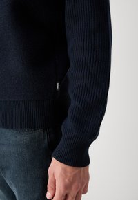 Maglione a maglia blu navy con polsini e orlo a coste. Presenta una texture morbida e un'etichetta con il marchio sul lato. Il dettaglio include una sezione del corpo liscia.