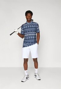 Kraťasové, námornícke golfové polo s vzorom palm, skombinované s bielymi šortkami. Model drží golfovú palicu, nosí biele č socks a čierne topánky.