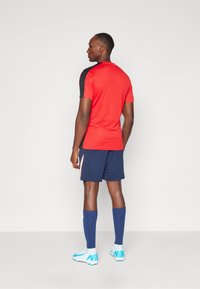 Camisa deportiva de manga corta roja y negra combinada con pantalones cortos azul marino, botas de fútbol azules y calcetines altos azules, mostrando un look deportivo.